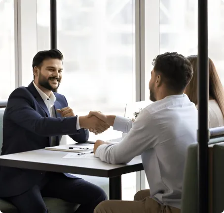 Two men in formal attire shaking hands in a bright office meeting room, with a window showing a city view.