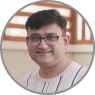 A smiling man with glasses, wearing a light-colored striped shirt, captured outdoors with a blurred wooden structure in the background.